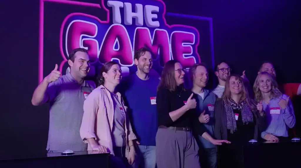 Gruppe von Spielern steht lachend vor dem leuchtenden „The Gameshow“-Logo in Hamburg – interaktives Live-Quiz für Teams, Freunde und Events